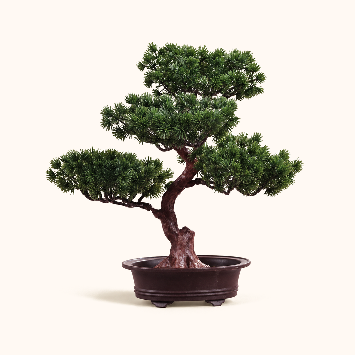 Small artificial pine bonsai tree with layered green foliage in a shallow dark oval pot on a cream background.